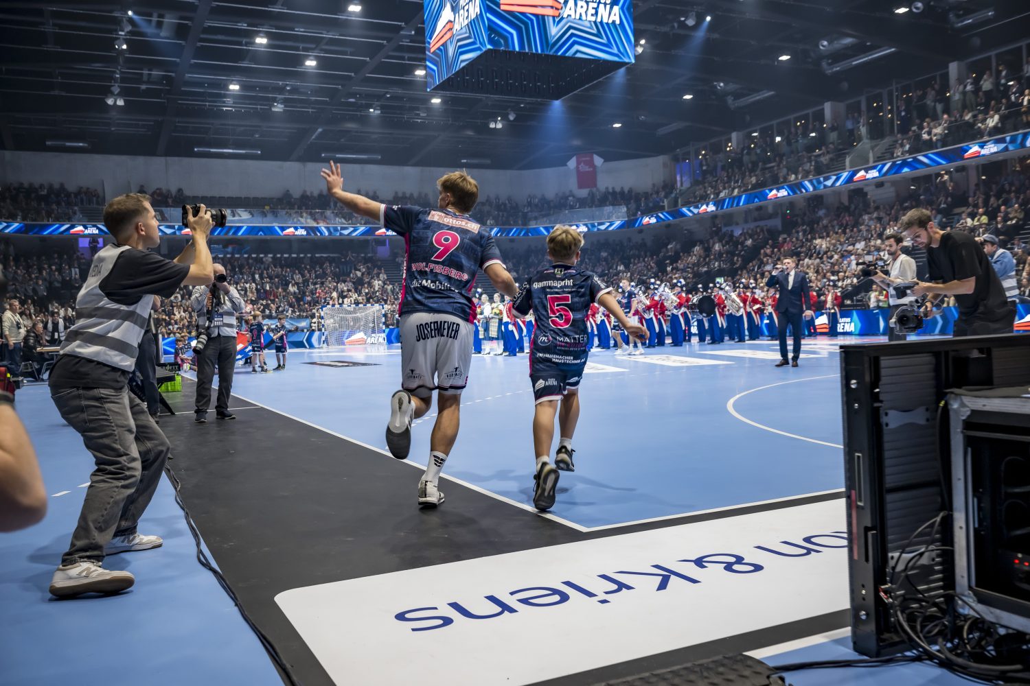 Zwei Handballerinnen des HC Kriens-Luzern mit den Nummern 9 und 5 laufen auf einem blauen Hallenplatz in der Schweiz auf eine jubelnde Menge zu. Fotografen und Veranstaltungsmitarbeiter halten den Moment fest, als sich ihre Mannschaft an der Seitenlinie aufstellt.