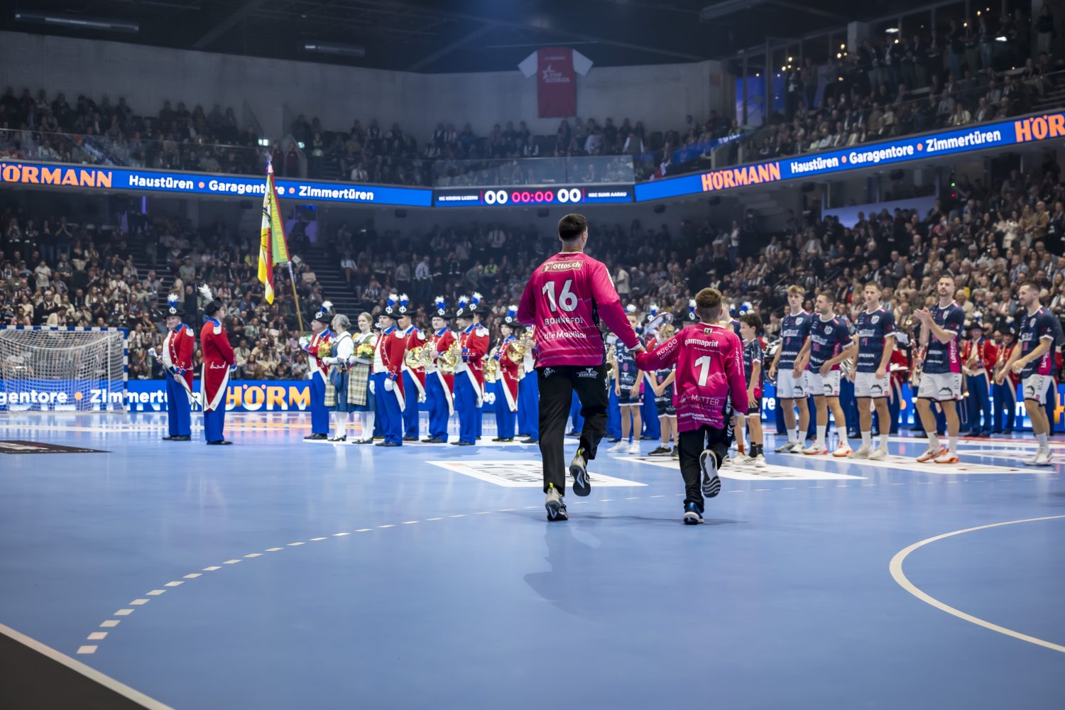 Zwei Handballer in rosa Trikots, ein Erwachsener und ein Kind, laufen Hand in Hand auf das hell erleuchtete Hallenfeld des HC Kriens-Luzern. Mannschaften, Schiedsrichter und eine Marschkapelle stehen in Formation, während ein volles Publikum von der Tribüne aus zusieht.