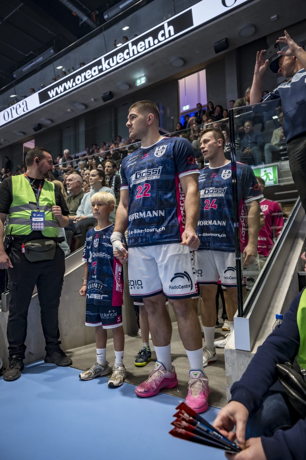 Ein Handballspieler im blau-rosa Trikot des HC Kriens-Luzern steht mit einem Kindermaskottchen und Mannschaftskameraden am Spielfeldrand und bereitet sich auf seinen Einsatz vor, während die Zuschauer in der Schweiz von der Tribüne aus zusehen.