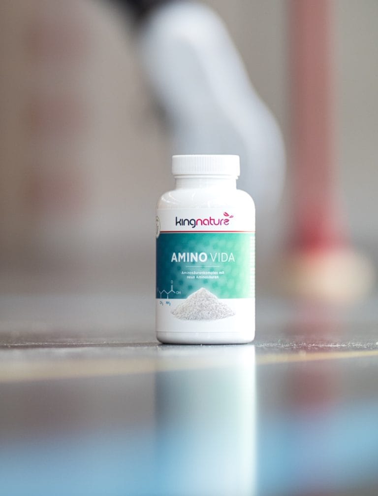 Eine Flasche des Nahrungsergänzungsmittels Kingnature Amino Vida steht auf einer reflektierenden Oberfläche mit unscharfem Hintergrund, was die unterstützende Wirkung von Amino Vida bei der Linderung von Schmerzen und Beschwerden hervorhebt. Ein weiß-rotes Objekt ist unscharf zu sehen.