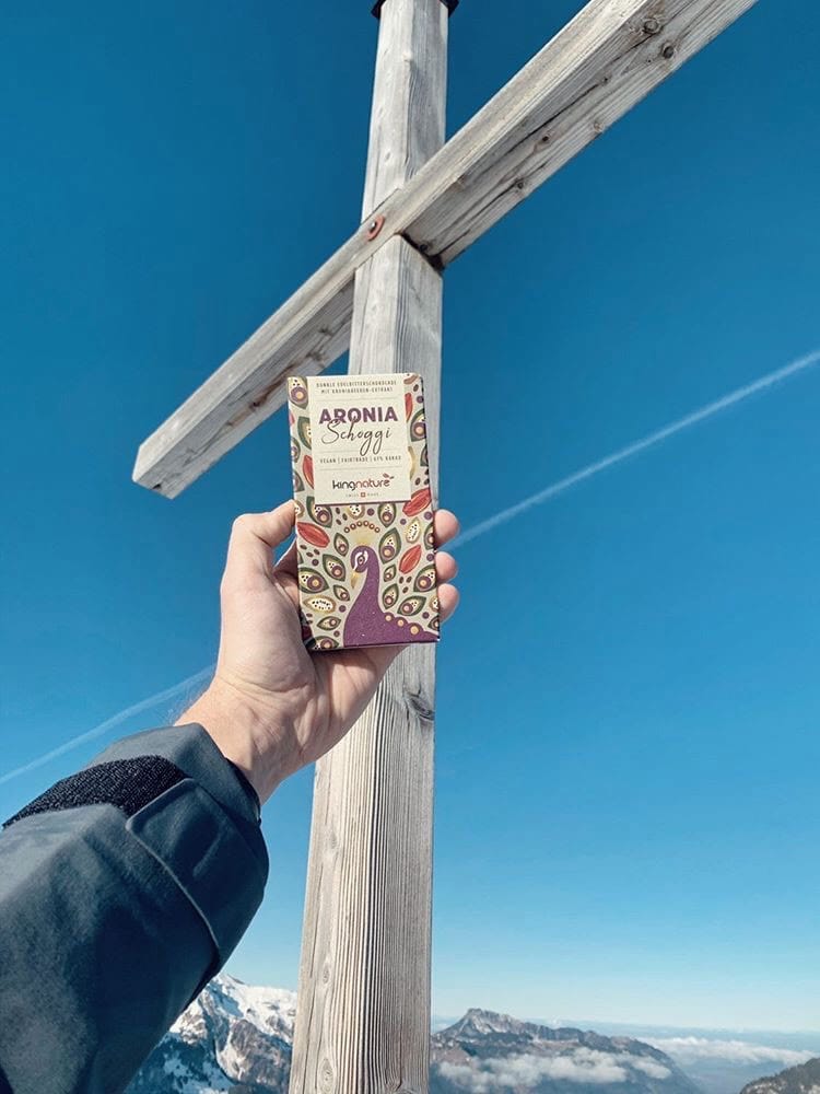 Eine Hand hält eine bunte Schachtel mit der Aufschrift „Aronia-Schokolade zum halben Preis“ vor einem großen Holzkreuz auf einem Berggipfel, im Hintergrund blauer Himmel und schneebedeckte Gipfel.
