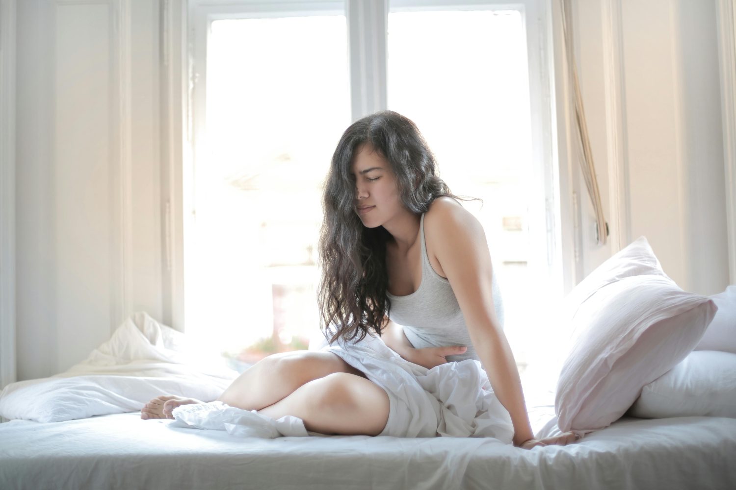 Eine Frau mit langen dunklen Haaren sitzt auf einem Bett vor einem hellen Fenster, hält sich den Bauch und scheint Schmerzen oder Unwohlsein zu haben - mögliche Symptome eines Vitamin-B12-Mangels. Sie ist von weißem Bettzeug und Kissen umgeben.