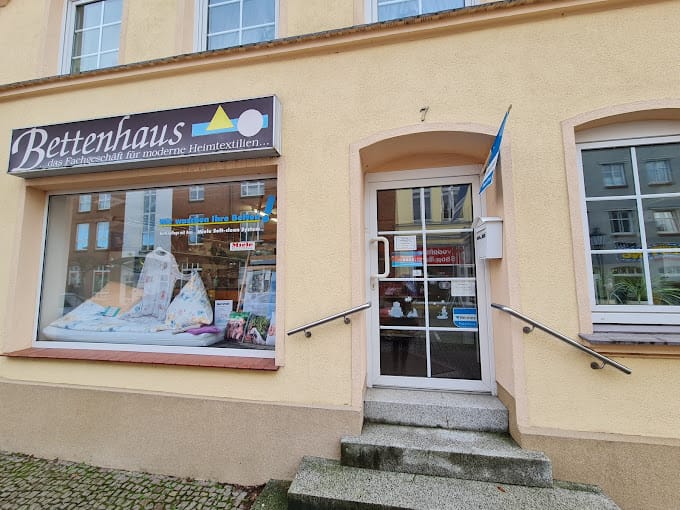 Ein Schaufenster mit einem großen Schaufenster, in dem Bettwaren und das Schild „Bettenhaus Heine“ ausgestellt sind. Neben dem Fenster befindet sich ein Eingang mit Glastür, Handläufen und Stufen, die hinaufführen. Die Fassade des Gebäudes ist hellgelb.