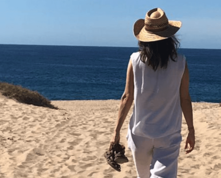 Eine Person in weißer Kleidung und Strohhut läuft barfuß an einem Sandstrand in Richtung des blauen Ozeans und hält dabei ihre Sandalen in einer Hand. Die Szene spiegelt die Ruhe der traditionellen chinesischen Gesundheitsvorsorge unter einem klaren, sonnigen Himmel wider.