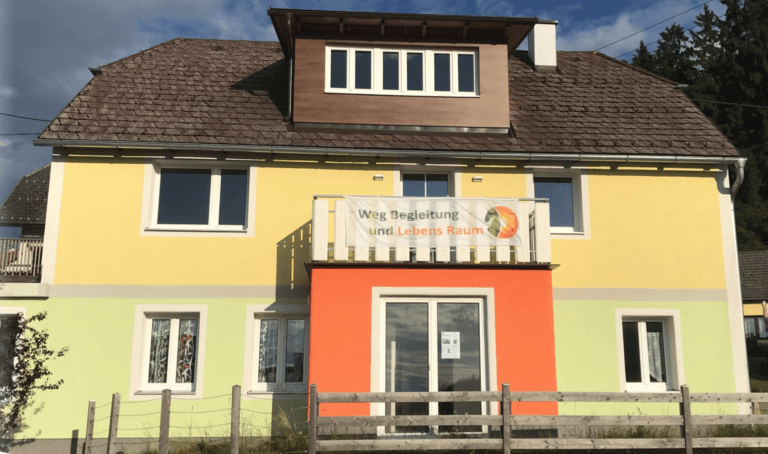 Ein zweistöckiges gelb-hellgrünes Gebäude mit Holzdach, großen Fenstern und einem Schild mit der Aufschrift „Weg Begleitung und Lebens Raum“ über dem Eingang. Brigitte Kofler bietet hier Coaching und Beratung an. Vor dem Gebäude befindet sich ein Holzzaun.