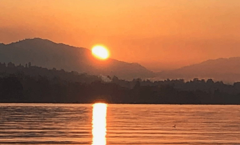 Eine goldene Sonne geht hinter fernen Bergen unter, wirft einen warmen orangefarbenen Schein über den Himmel und spiegelt sich im ruhigen Wasser mit sanften Kräuselungen und einer schwachen Silhouette von Bäumen – und ruft so die ruhige Atmosphäre von Jambor Hypnose hervor.