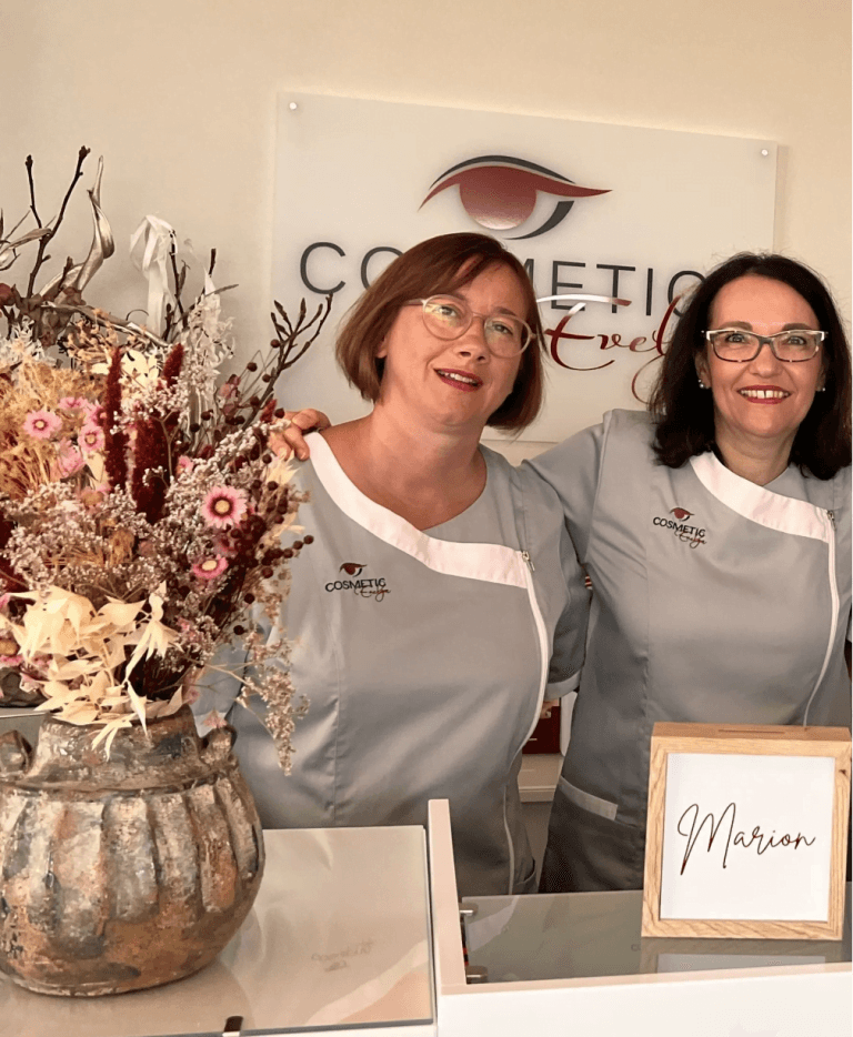 Zwei Frauen in passenden grauen Uniformen stehen lächelnd an einem Kosmetikempfangstresen mit einem Schild mit der Aufschrift „Marion“ und einer Vase mit Trockenblumen. An der Wand hinter ihnen prangt ein Logo mit einem stilisierten Auge, das dem Raum einen Evelyn-Touch verleiht.