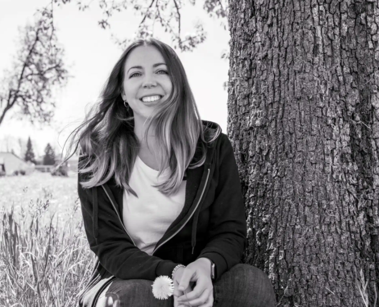 Eine junge Frau mit langen Haaren sitzt lächelnd neben einem Baum auf einer Wiese. Sie trägt ein helles T-Shirt und eine dunkle Jacke und hält eine kleine Blume in der Hand. Stephanie Iaccarino hält diesen von Naturheilkunde inspirierten Schwarz-Weiß-Moment fest.