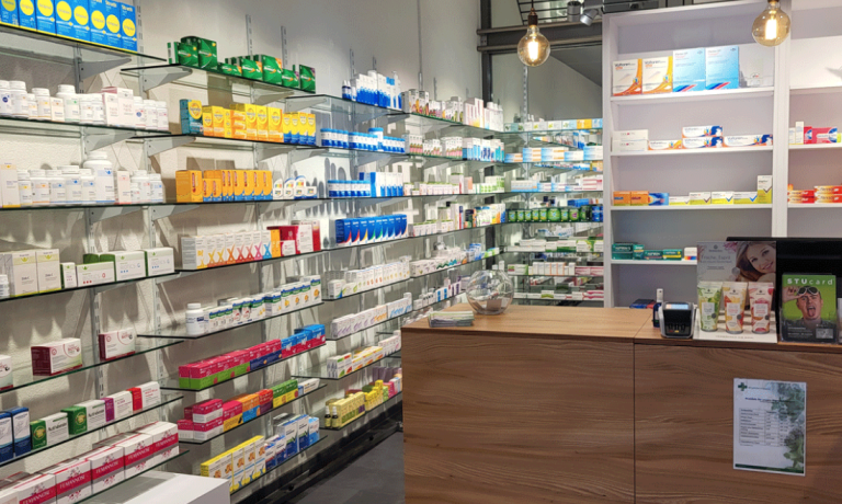 Ein modernes Interieur einer Pill Apotheke mit Glasregalen, auf denen verschiedene Schachteln ausgestellt sind, einem Holztresen mit Zahlungsterminal und heller Beleuchtung.