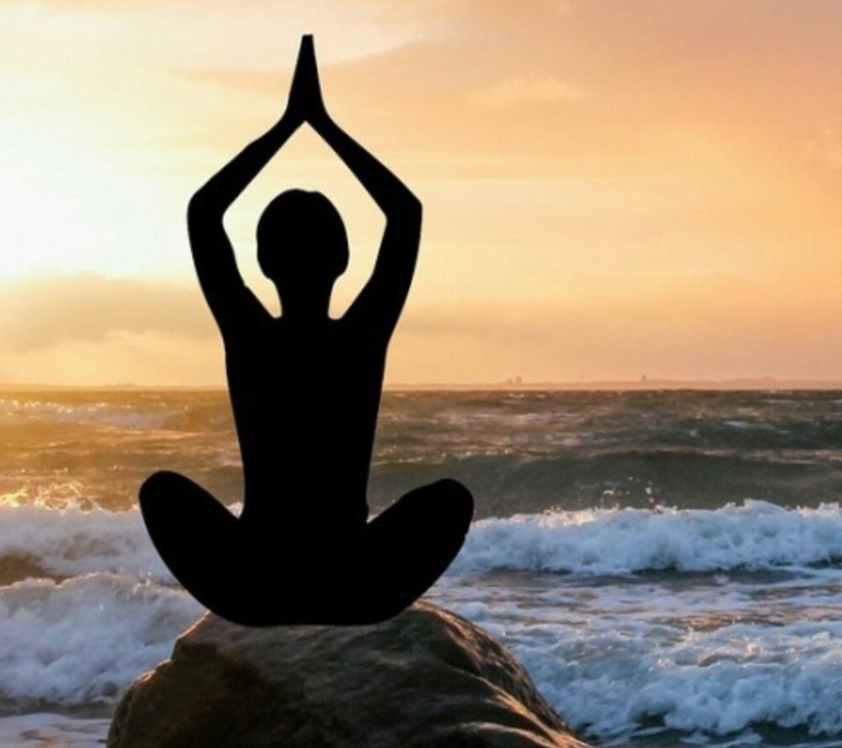 Eine Person sitzt im Schneidersitz auf einem Felsen am Meer und hält die Hände in einer Yoga-Pose über dem Kopf. In der Nähe brechen Wellen, während hinter ihnen ein warmer, goldener Sonnenuntergang leuchtet und den friedlichen Geist von Craniosa und Andrea Kübler heraufbeschwört.