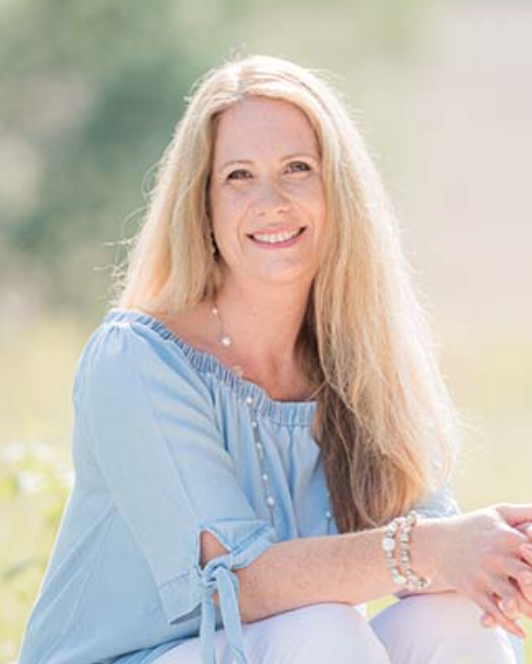 Miriam Meister, mit langen blonden Haaren, trägt ein hellblaues schulterfreies Top und eine weiße Hose. Sie sitzt an einem sonnigen Tag im Freien und lächelt in die Kamera. Der weiche, unscharfe Hintergrund lässt Grün erahnen – eine einladende Szene für AT-Therapie und Ernährung.