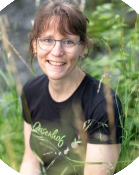 Therese Berger, mit Brille und schwarzem T-Shirt, lächelt in die Kamera, während sie draußen im hohen grünen Gras des Oasenhofs sitzt.