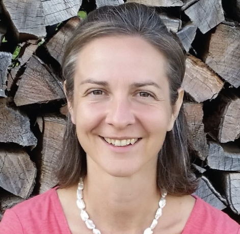 Manuela Hofstetter, mit glattem braunem Haar, einem rosa Oberteil und einer weißen Perlenkette, lächelt vor einem Stapel Brennholz in der Praxis für Lebendigkeit.