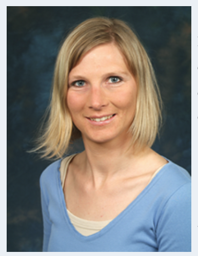 Eine Frau mit glattem blondem Haar und blauen Augen, die ein hellblaues Top über einem beigen Hemd trägt, lächelt vor einem dunkelblau gesprenkelten Hintergrund in die Kamera und strahlt Selbstvertrauen aus.