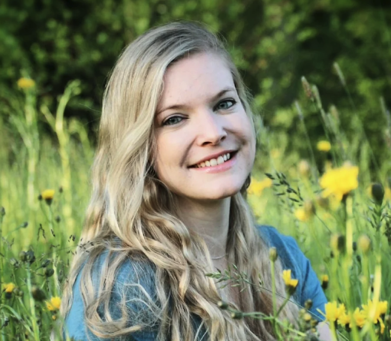 Eine junge Frau mit langen blonden Haaren lächelt, während sie auf einem Feld mit hohem Gras und gelben Wildblumen sitzt, umgeben von üppigem grünem Laub.