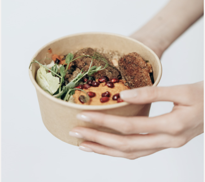 Vor einem schlichten Hintergrund hält eine Person eine braune Papierschüssel mit gemischtem Salat, Falafel und proteinreichem Hummus, garniert mit Granatapfelkernen und anderen frischen Zutaten.