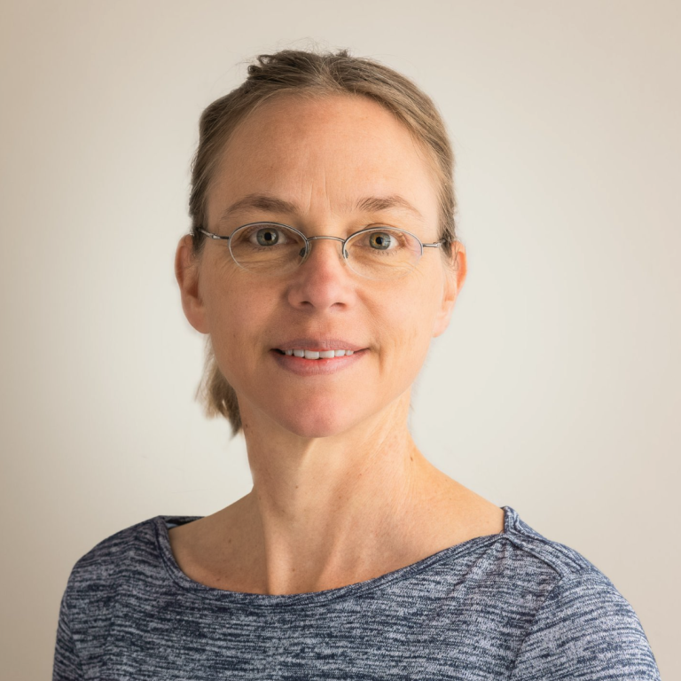 Eine Frau mit zurückgekämmtem hellbraunem Haar, Brille und blau gemustertem Oberteil lächelt vor einem schlichten, hellen Hintergrund sanft in die Kamera und hebt so positive Verkaufspunkte hervor.
