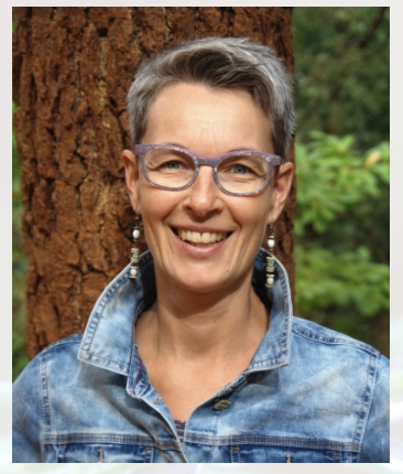 Eine lächelnde Person mit kurzen grauen Haaren und Brille steht vor einem Baum. Sie trägt eine blaue Jeansjacke und lange Ohrringe. Im Hintergrund sind grüne Blätter und Verkaufspunkte zu sehen.