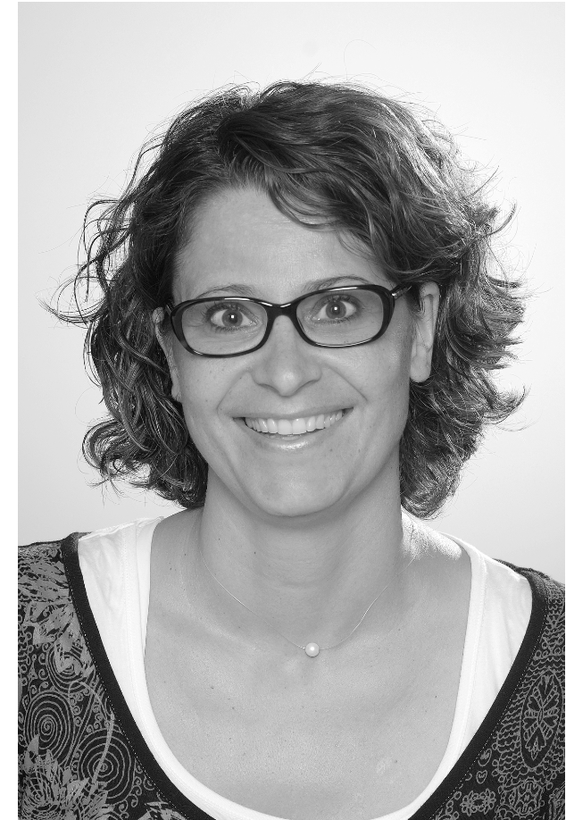 Schwarz-Weiß-Porträt einer Frau mit kurzen, lockigen Haaren, Brille, schlichter Halskette und gemusterter Kleidung. Sie lächelt in die Kamera. Das Bild eignet sich gut für Werbematerialien zur Hervorhebung von Verkaufspunkten.