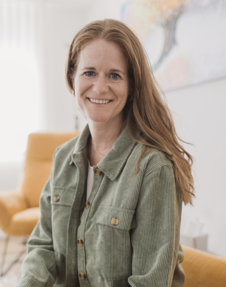 A person with long, reddish-brown hair and a green corduroy shirt smiles at the camera. They are sitting in a light, airy room with a yellow chair and abstract artwork in the background.