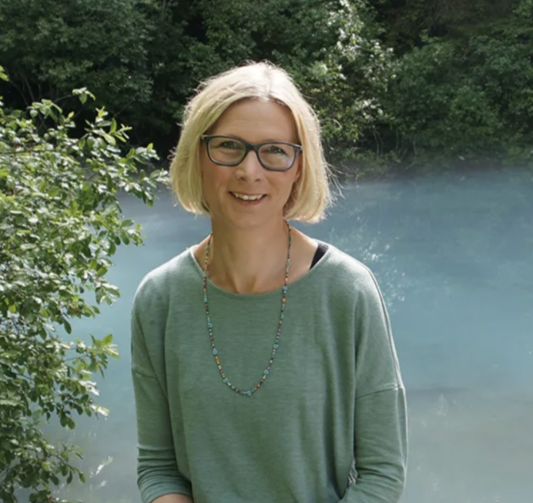 Milena Matter-Meisser, Fachärztin für Sportpsychologie, steht mit kurzen blonden Haaren und Brille im Freien, trägt ein hellgrünes Oberteil und eine Perlenkette, umrahmt von dichtem grünen Laub und blauem Wasser.