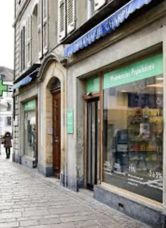 Ein Apothekenladen mit großen Fenstern und grünen Schildern auf einer Kopfsteinpflasterstraße in einer europäischen Stadt; ein automatisch gespeicherter Entwurf des täglichen Lebens, mit einer Person, die in der Ferne geht.