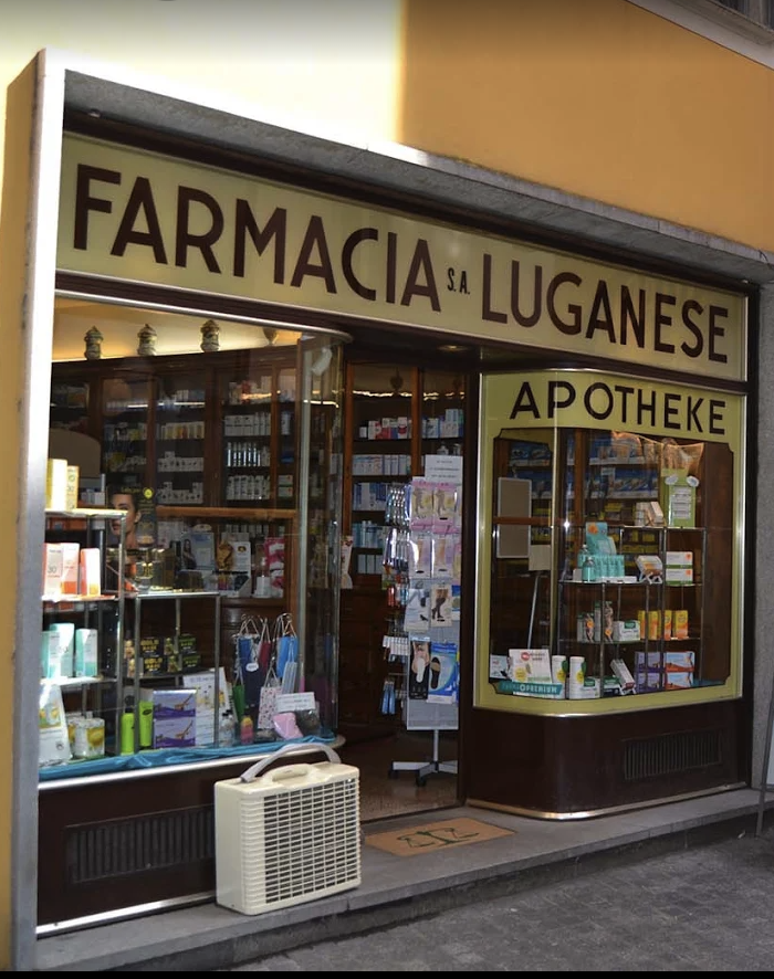 Die Schaufensterfront einer Apotheke mit dem Namen "Farmacia Luganese" und der Aufschrift "Apotheke" auf dem Schild. Im Schaufenster werden verschiedene Gesundheits- und Schönheitsprodukte angeboten, und in der Nähe der Klimaanlage am Eingang erscheint ein Hinweis auf einen automatisch gespeicherten Entwurf.