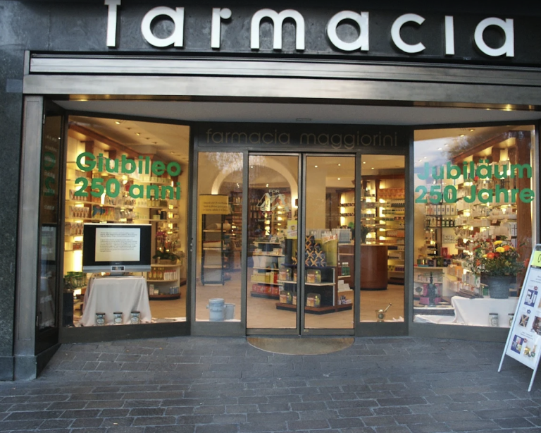 Schaufenster einer Apotheke namens "Farmacia Maggiorini" mit großen Glastüren, Regalen mit Produkten im Inneren und grünen Schildern in italienischer und deutscher Sprache zum 250-jährigen Jubiläum - ein automatisch gespeicherter Entwurf von Tradition und Sorgfalt.