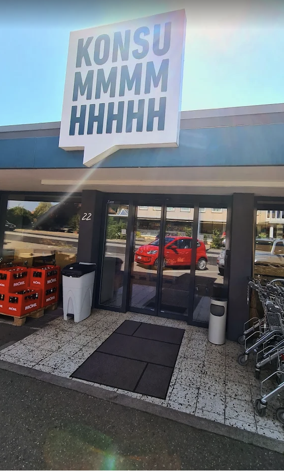 Der Eingang eines Lebensmittelladens mit einem großen Schild, auf dem KONSUMMMMMHHHHH steht, wirkt wie ein automatisch gespeicherter Entwurf. Auf der rechten Seite stehen Einkaufswagen, links neben der Tür stapeln sich rote Kisten, und draußen parkt ein rotes Auto.