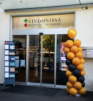 Der Eingangsbereich der Rotpunkt Apotheke in Vindonissa mit Glastüren, einem Schild darüber, einer Säule aus schwarzen und orangefarbenen Luftballons sowie einem Ausstellungsständer mit Broschüren neben der Tür – ein automatisch gespeicherter Entwurf zur Begrüßung der Besucher.