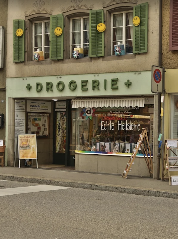 Ein Ladenlokal mit der Aufschrift „Drogerie“, grünen Fensterläden, gelben Smileys und verschiedenen Schildern vor dem Eingang – darunter ein Prospektständer mit der Aufschrift „Automatisch gespeicherter Entwurf“ – sticht durch seine beigen Wände und großen Fenster hervor.