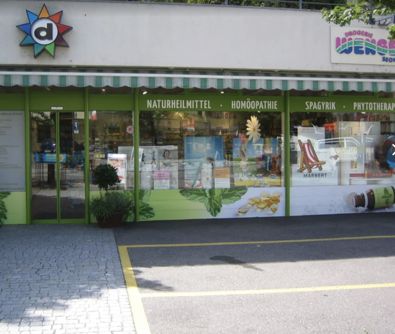 Ein Ladenlokal mit grün gerahmten Fenstern und einer gestreiften Markise, auf der Begriffe wie „Naturheilmittel“, „Homöopathie“ und „Phytotherapie“ zu lesen sind. Neben Pflanzen- und Kräuterbildern ist im Schaufenster ein „automatisch gespeicherter Entwurf“ zu sehen, über der Tür befindet sich ein Apothekenlogo.