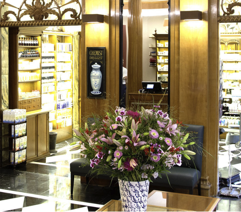 Ein großer Strauß aus rosa und violetten Blumen in einer dekorativen Vase steht auf einem Holztisch in der luxuriösen St. Peter Apotheke in Zürich, einer hell beleuchteten Apotheke mit kunstvollen Holzvertäfelungen und Regalen voller Produkte.