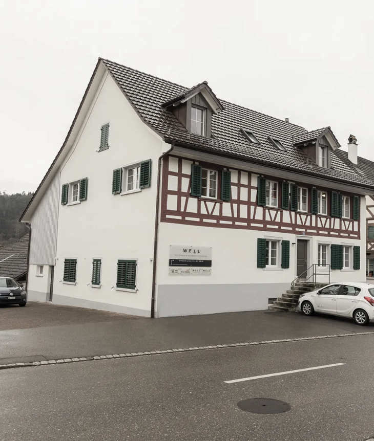 An einer abschüssigen Straße steht ein traditionelles europäisches Gebäude mit weißen Wänden, grünen Fensterläden und Fachwerk. Daneben sind mehrere Autos geparkt, und in der Nähe der Eingangstreppe ist ein Schild mit der Aufschrift „WELL“ zu sehen – wie ein automatisch gespeicherter Entwurf der Geschichte.