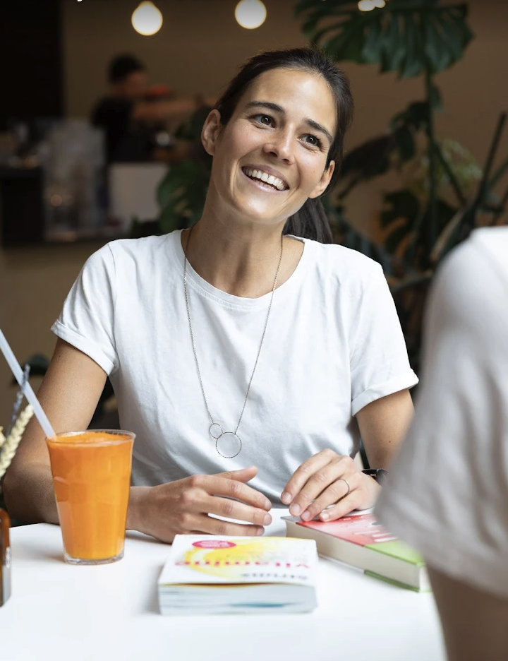 Eine lächelnde Frau in einem weißen T-Shirt sitzt an einem Tisch mit Büchern und einem Glas Orangensaft und unterhält sich in einem hellen, gemütlichen Café. Große grüne Pflanzen rahmen die Szene ein – wie ein automatisch gespeicherter Entwurf eines perfekten Morgens.