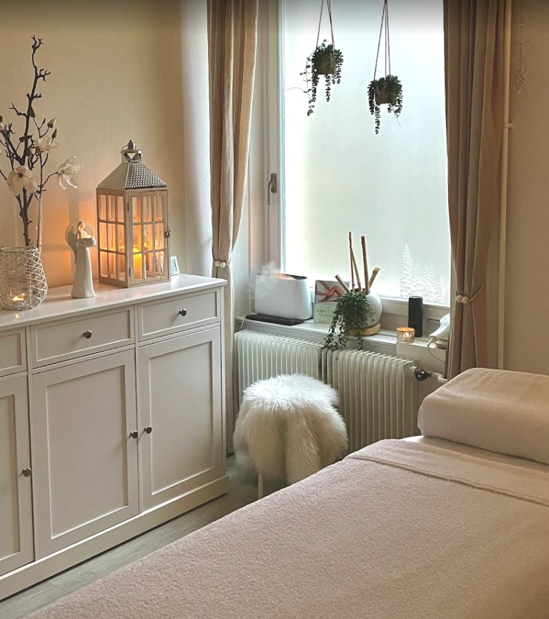 Ein gemütlicher Wellnessraum mit einer Massageliege, weicher beiger Bettwäsche, einem flauschigen weißen Hocker, Kerzen und Topfpflanzen auf der Fensterbank empfängt die Gäste in einer Atmosphäre, die so ruhig und beruhigend ist wie ein automatisch gespeicherter Entwurf – mühelos und gelassen.