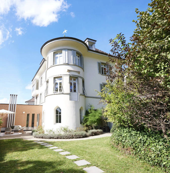 Ein weißes, dreistöckiges Haus mit abgerundeten Ecken und blauen Fensterläden steht neben einem üppigen Garten mit einem Steinweg unter strahlend blauem Himmel – wie ein automatisch gespeicherter Entwurf eines friedlichen Rückzugsortes.