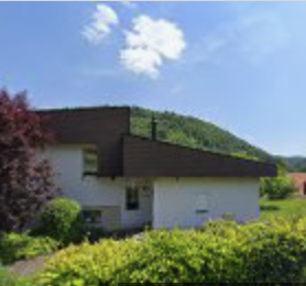 Ein modernes Haus mit dunkelbraunen und weißen Außenwänden, umgeben von grünen Sträuchern und Bäumen, steht auf einem grasbewachsenen Hügel unter blauem Himmel – ein automatisch gespeicherter Entwurf zeitgenössischen Designs im Einklang mit der Natur.
