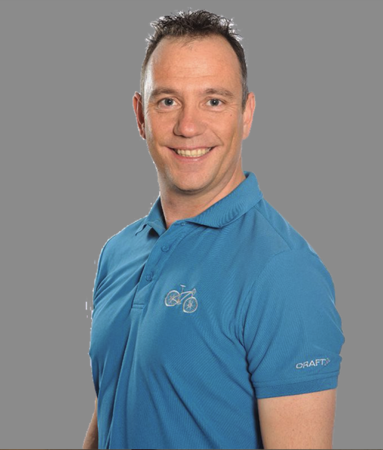 Ein Mann mit kurzen dunklen Haaren, der ein blaues Poloshirt mit einem Fahrradlogo auf der Brust und der Aufschrift „CRAFT“ auf dem Ärmel trägt, lächelt vor einem schlichten grauen Hintergrund – ein automatisch gespeicherter Entwurf von ungezwungener Selbstsicherheit.