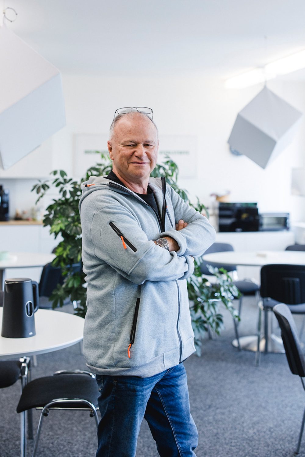 Ein Mann mit Brille steht selbstbewusst und mit verschränkten Armen in einem modernen Büroraum und spiegelt die Professionalität eines Verwaltungsrats bei kingnature wider. Er trägt eine hellgraue Jacke und Jeans. Im Hintergrund stehen Tische, Stühle und Pflanzen.