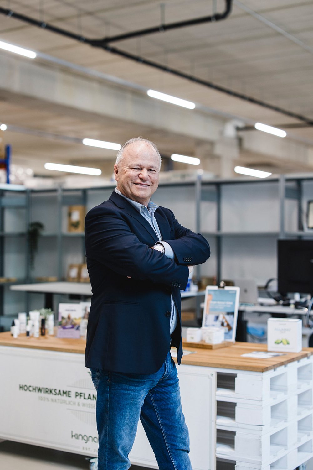 Ein lächelnder älterer Herr in dunklem Blazer und Jeans steht selbstbewusst mit verschränkten Armen in einem modernen, hellen Büroraum. Im Hintergrund sind Regale und kingnature-Produkte zu sehen, die seine Rolle im Verwaltungsrat widerspiegeln.