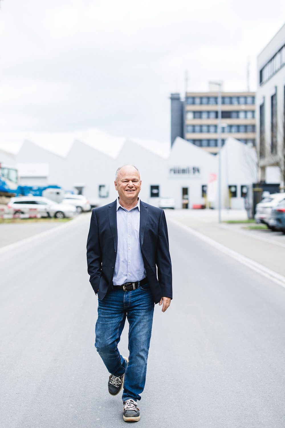 Ein Mann in marineblauem Blazer, hellem Hemd und Jeans geht selbstbewusst eine leere Stadtstraße entlang, die Hände in den Taschen, lächelnd – vielleicht auf dem Weg zu einer Verwaltungsratssitzung bei kingnature. Im Hintergrund sind moderne Gebäude und parkende Autos zu sehen.