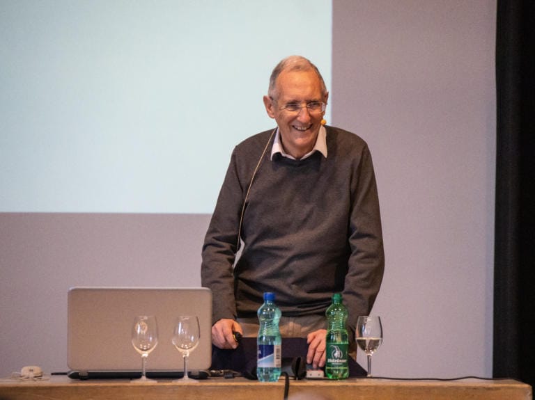 Ein älterer Mann mit Brille steht lächelnd hinter einem Tisch mit einem Laptop, vier leeren Weingläsern und zwei Wasserflaschen und unterhält sein Publikum vor einer leeren Projektionsfläche mit einer Präsentation zum Thema Nachhaltigkeit.