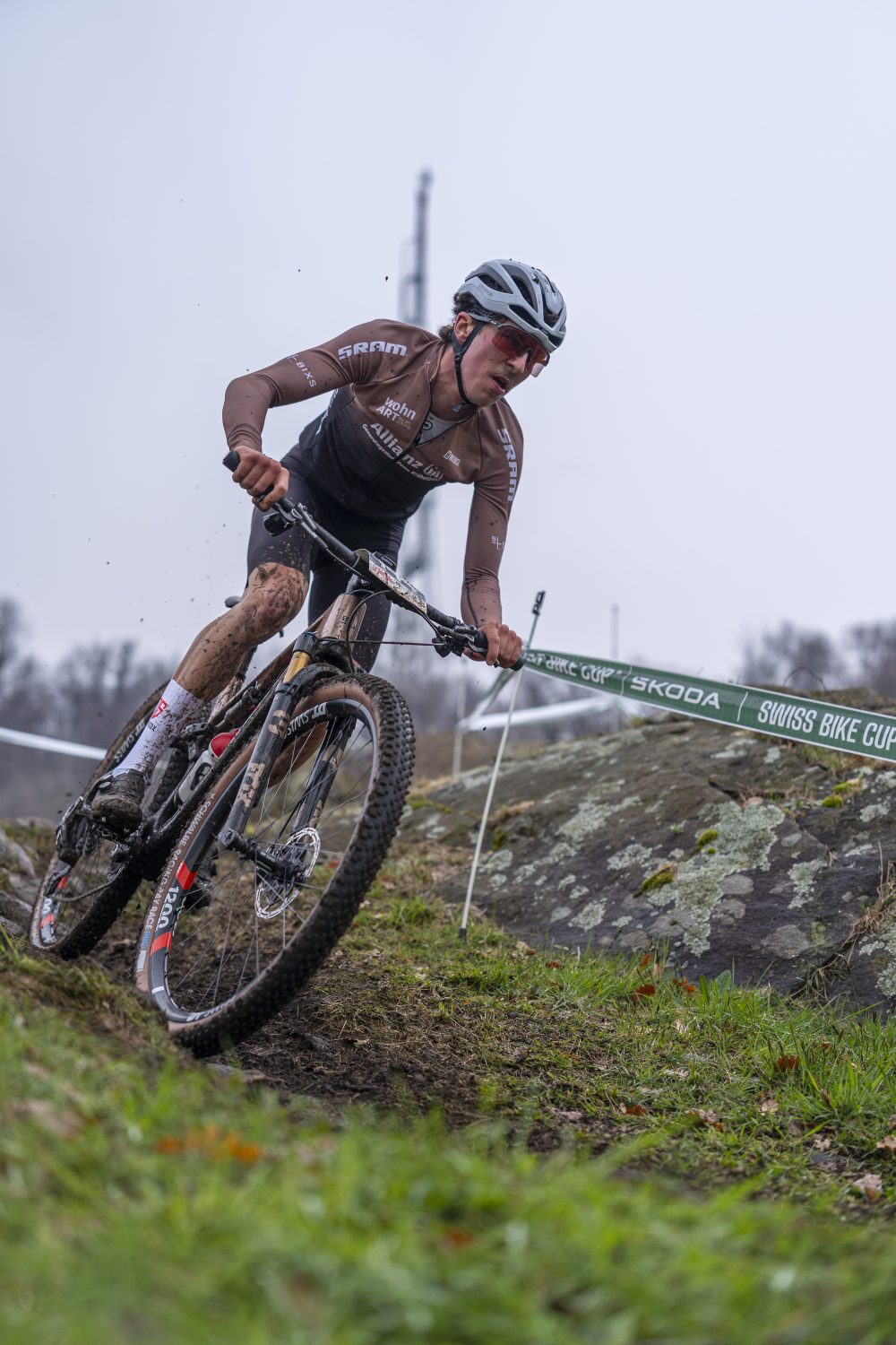 Timon Minder, der einen Helm und ein braunes Trikot trägt, fährt mit seinem Mountainbike auf einem schlammigen, grasbewachsenen Weg und neigt sich in eine Kurve. Die Umgebung besteht aus Felsen, einer grünen Barriere und einem bedeckten Himmel.