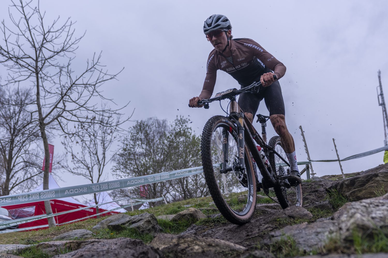 Timon Minder, der einen Helm und ein braunes Trikot trägt, fährt während eines Rennens an einem bewölkten, nassen Tag mit dem Mountainbike über felsiges, schlammiges Gelände. Kahle Bäume, Banner und Absperrungen füllen den Hintergrund.