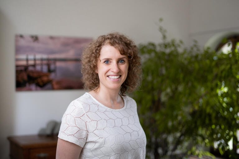 Eine Frau mit lockigem hellbraunem Haar und einem weißen gemusterten Hemd steht lächelnd in der Naturheilpraxis TEN. Eine grüne Pflanze und ein Landschaftsfoto schmücken den Hintergrund.