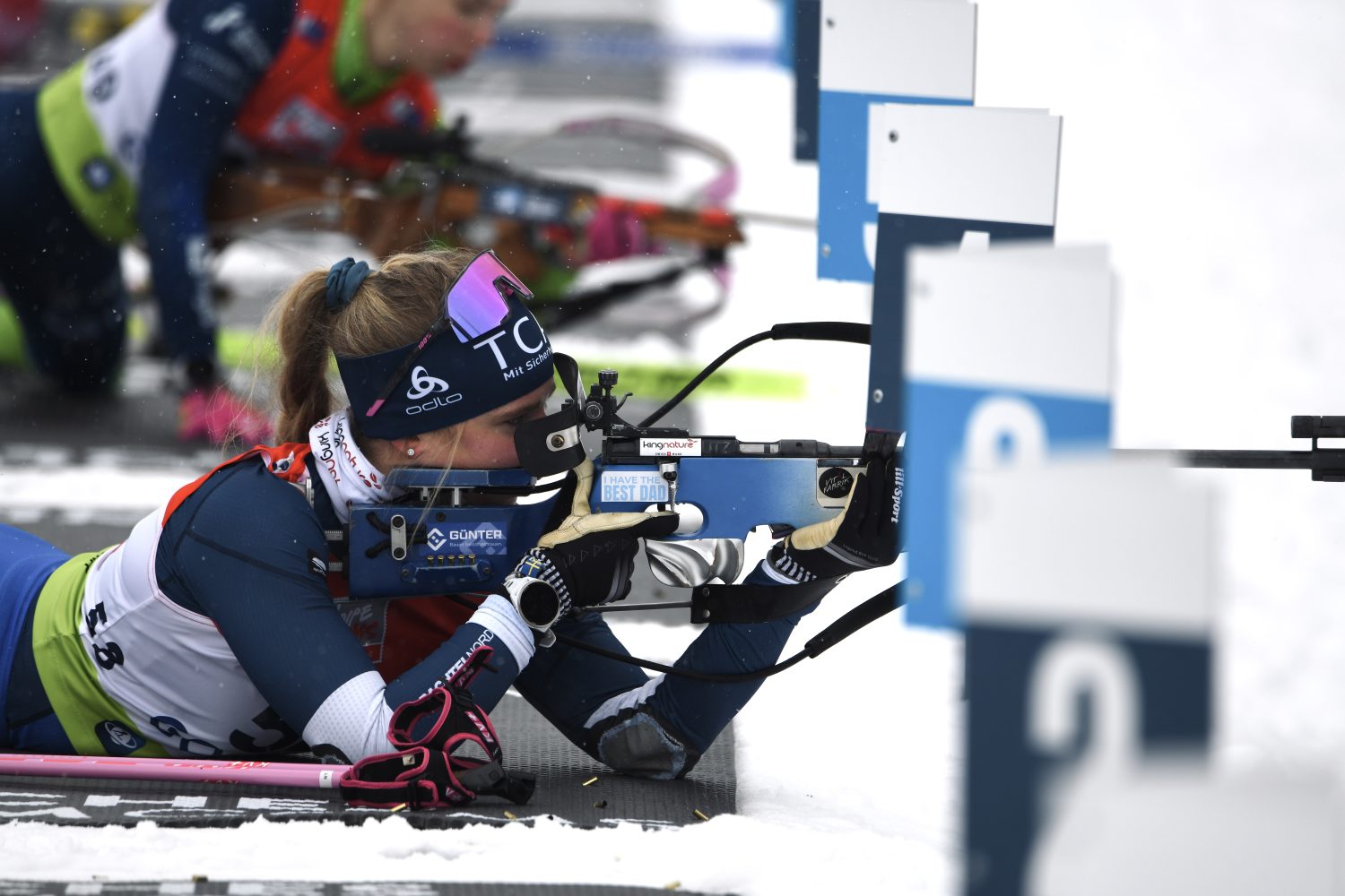 Die Biathletin Lisa Naegeli in Wintersportkleidung zielt auf ihr Gewehr, während sie während eines Biathlon-Wettkampfs auf dem Schießstand liegt. Der Schnee bedeckt den Boden, während sich andere Athleten im Hintergrund auf das Schießen vorbereiten.