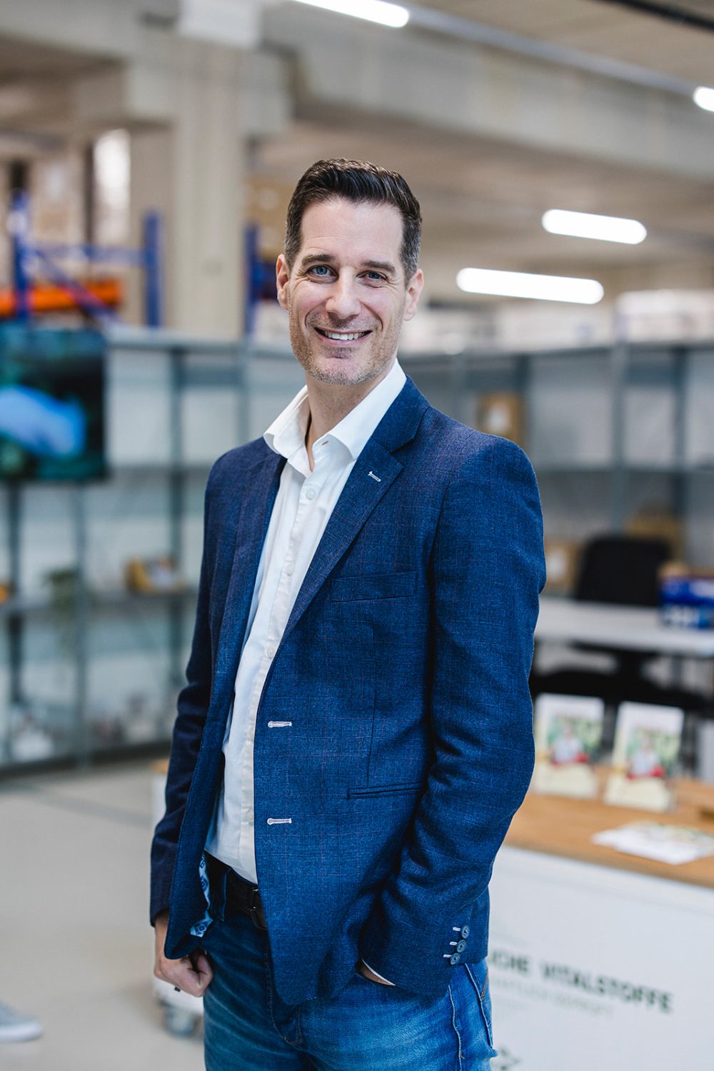 Ein Verwaltungsratsmitglied in einem blauen Blazer und Jeans steht lächelnd in einem modernen Büro oder Arbeitsbereich mit Regalen und einem Schreibtisch und spiegelt damit das professionelle Ethos von kingnature wider.