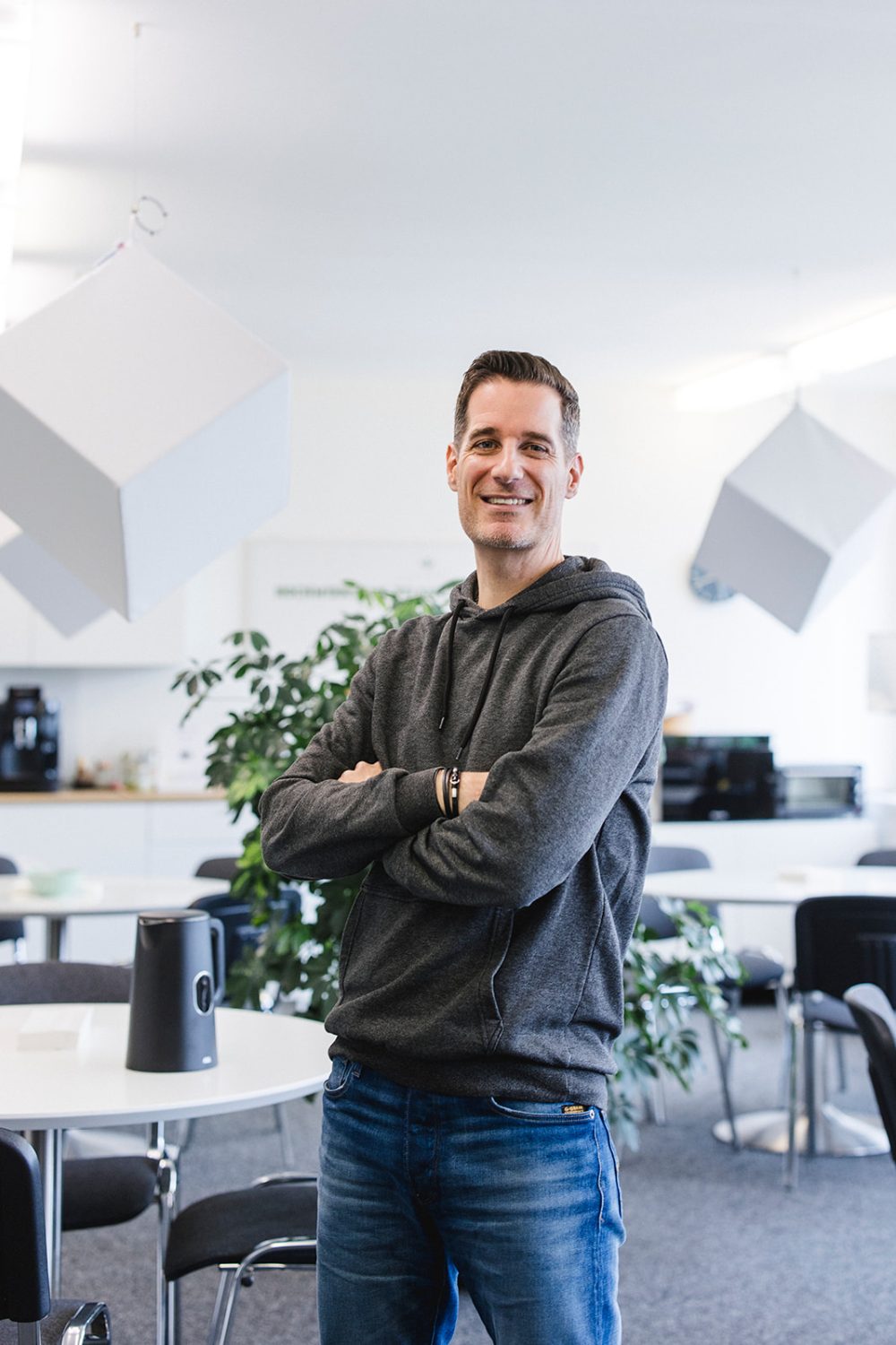 Ein Mann in einem grauen Kapuzenpullover und Jeans steht lächelnd und mit verschränkten Armen in einem modernen, hellen Büroraum mit weißen Tischen, Stühlen und geometrischen Hängedekorationen – ein Spiegelbild der dynamischen Energie des kingnature Verwaltungsrats.