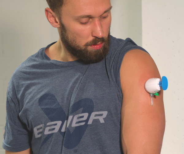 Ein bärtiger Mann in einem blauen T-Shirt schaut auf seinen Oberarm, an dem ein medizinischer Sensor TAP-Blut-Test Blei-Check angebracht ist.